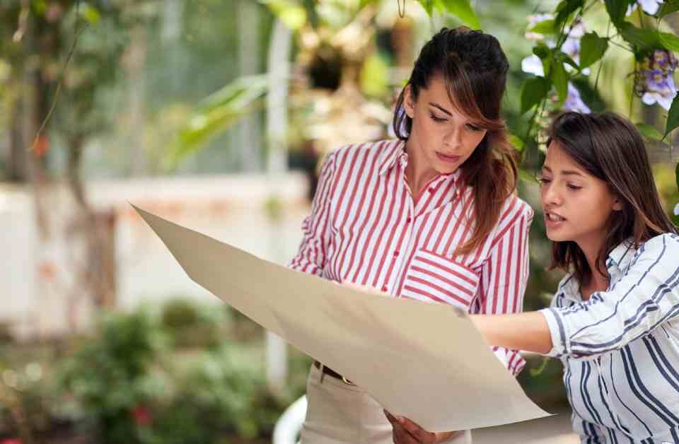 Two women stand in a garden. They are looking at a paper design plan, and one is pointing out details to the other.